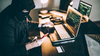 Hacker using a phone and pc while wearing a ski mask. 