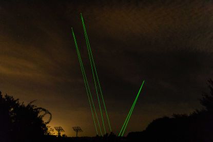Night image, dark cloudy sky, three sets of green laser beams projected up to the sky from ground level, silhouette of people, trees and two aerials
