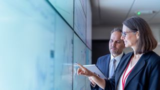 Two tech leaders talking and gesturing towards a large screen on the wall. The image is shot in close-up.