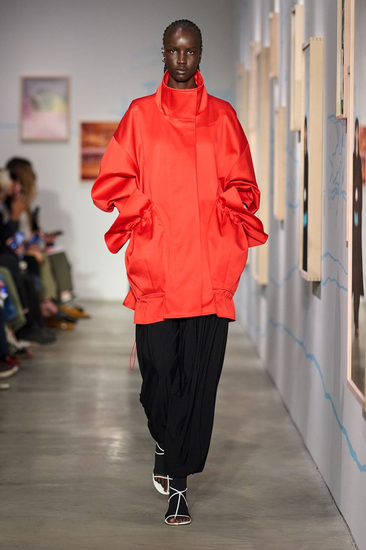 An Ashlyn model wearing an oversize bright red funnel-neck jacket styled with black wide-leg trousers, black tights, and white T-strap sandals.