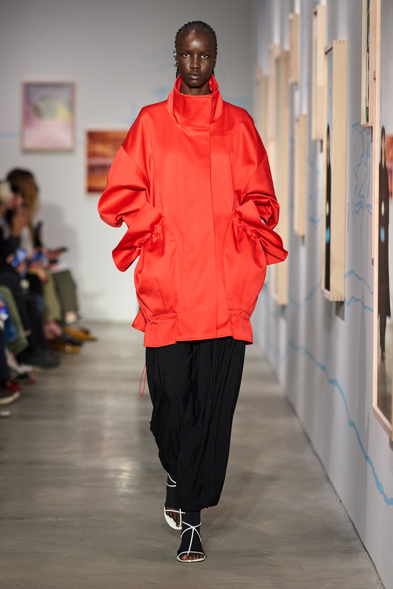 An Ashlyn model wearing an oversize bright red funnel-neck jacket styled with black wide-leg trousers, black tights, and white T-strap sandals. 