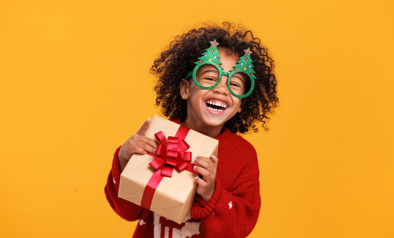 A boy wearing funny glasses in form of Christmas trees laughing while holding xmas gift box, having fun while standing yellow background