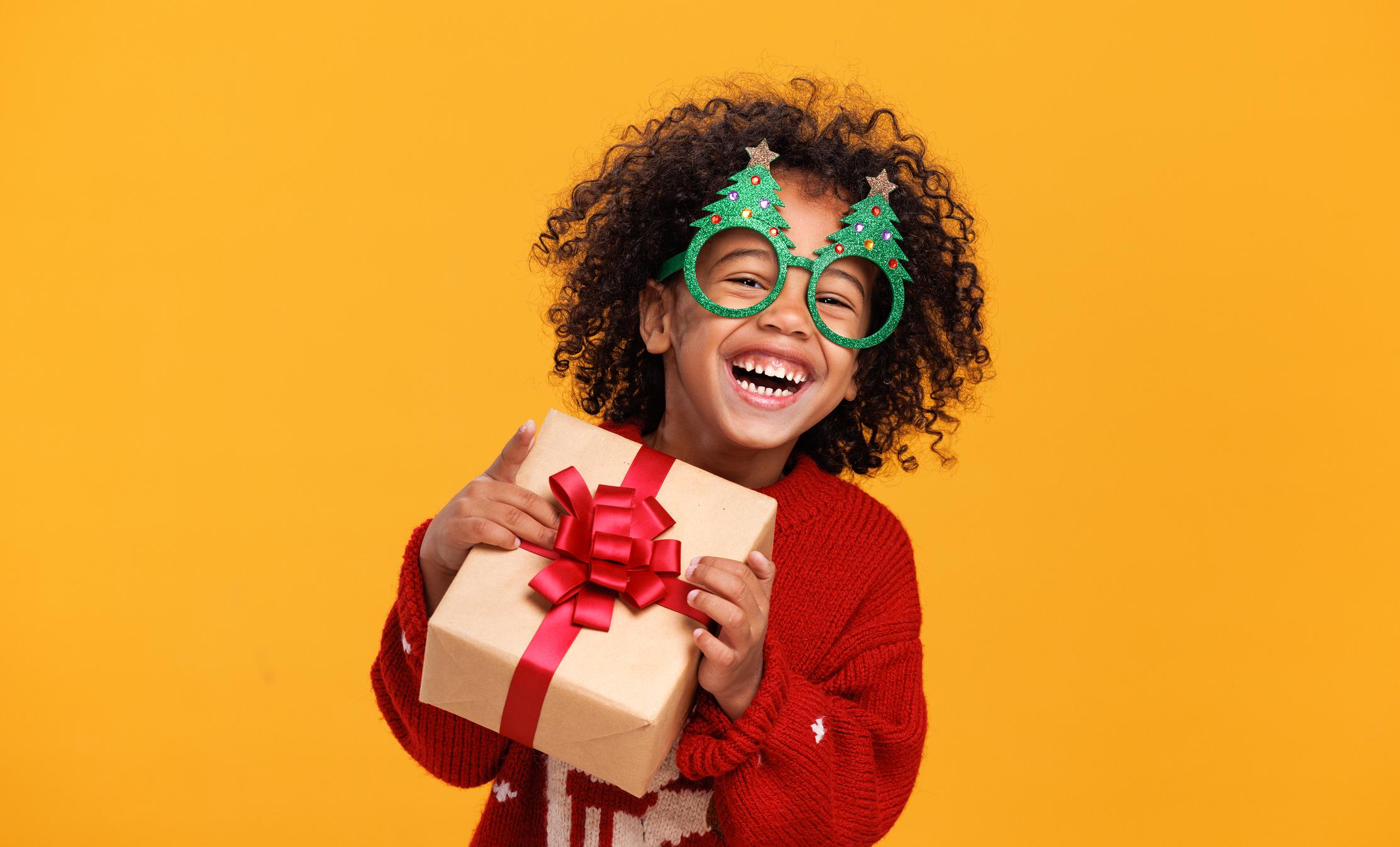  A boy wearing funny glasses in form of Christmas trees laughing while holding xmas gift box, having fun while standing yellow background 
