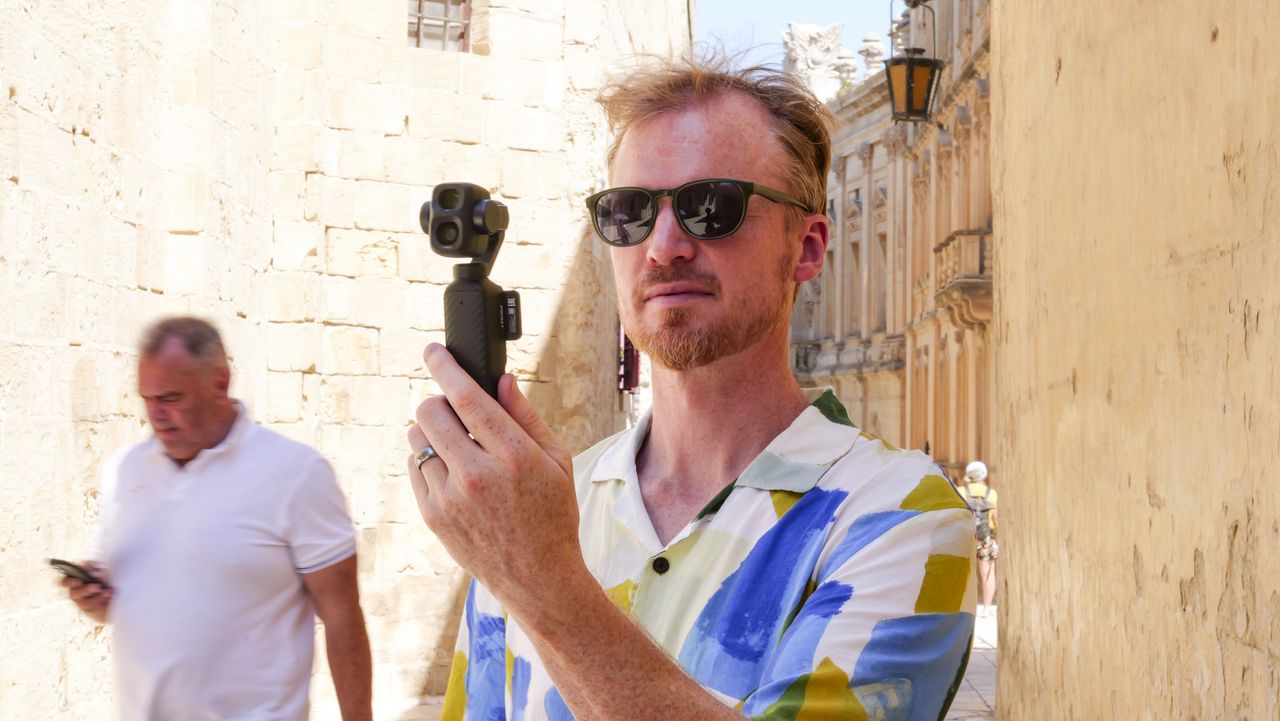 Man in bright summer short and shades, in an old town on a sunny day, holding the DJI Osmo Pocket 4P vlogging camera