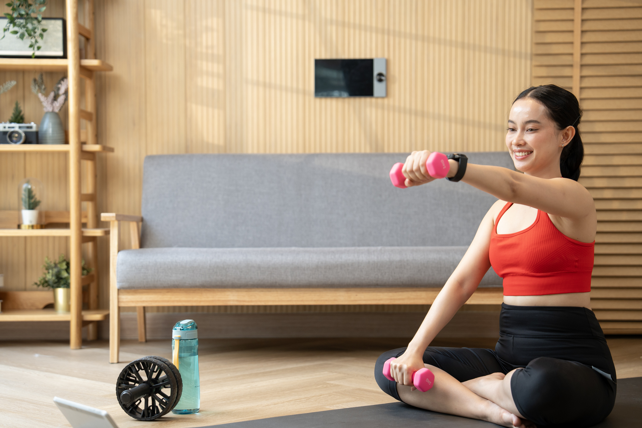 A woman works out from her home with pink dumbbells, an ab whee and a water bottle