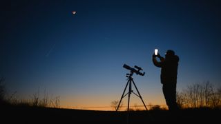 A man standing next a telescope and holding up his smartphone to the moon and night sky