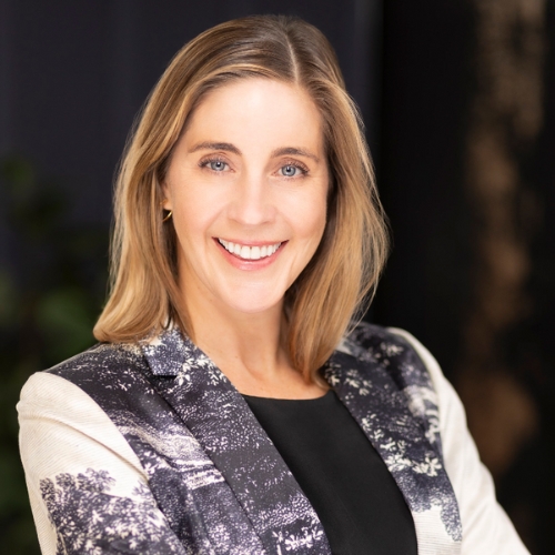 Image of a woman in a black shirt and a blue and white patterned blazer standing in front of a dark background.