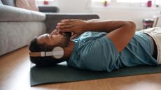 Man lying on his back on a yoga mat in a domestic setting. He is wearing headphones and has his eyes closed. His palms are together with his index fingers resting on his mouth.