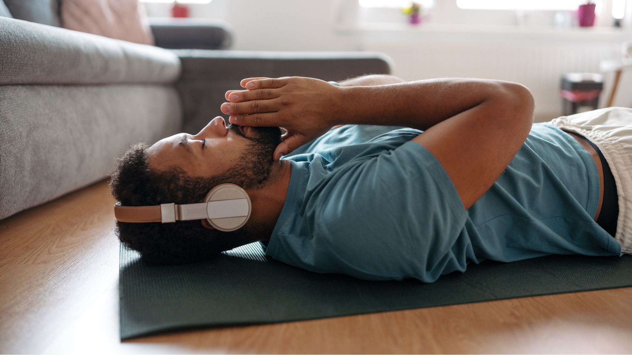 Man lying on his back on a yoga mat in a domestic setting. He is wearing headphones and has his eyes closed. His palms are together with his index fingers resting on his mouth.