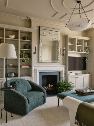 Image of a beige and off-white living room with a black and marble fireplace. There are built-in bookshelves on either side of the fireplace and a mirror hanging over it. In the sitting area there is a phthalo green boucle accent chair, a phthalo green ottoman, and a white sofa partially in view. There is a black and white floor lamp with a built-in tray.