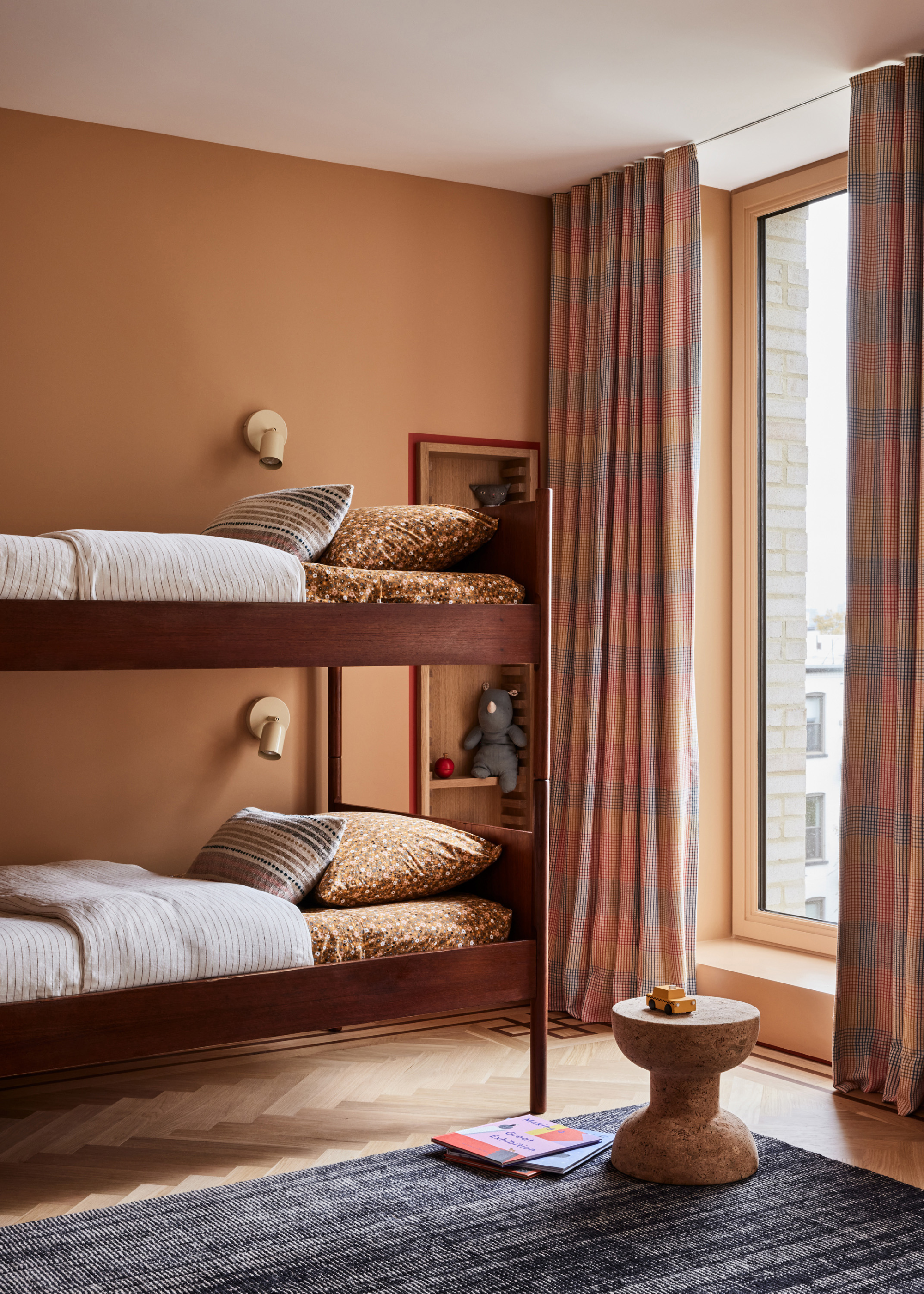 A kids bedroom with a cork stool, a bunk bed, beige wall sconces, a storage nook, and patterned curtains