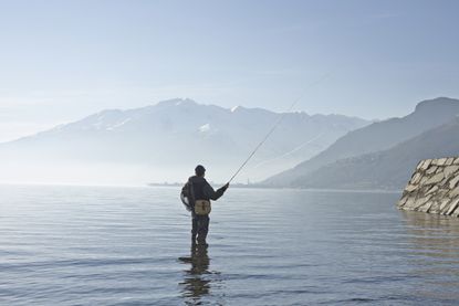 A man fishing in waders with mountains in the background, alone. 