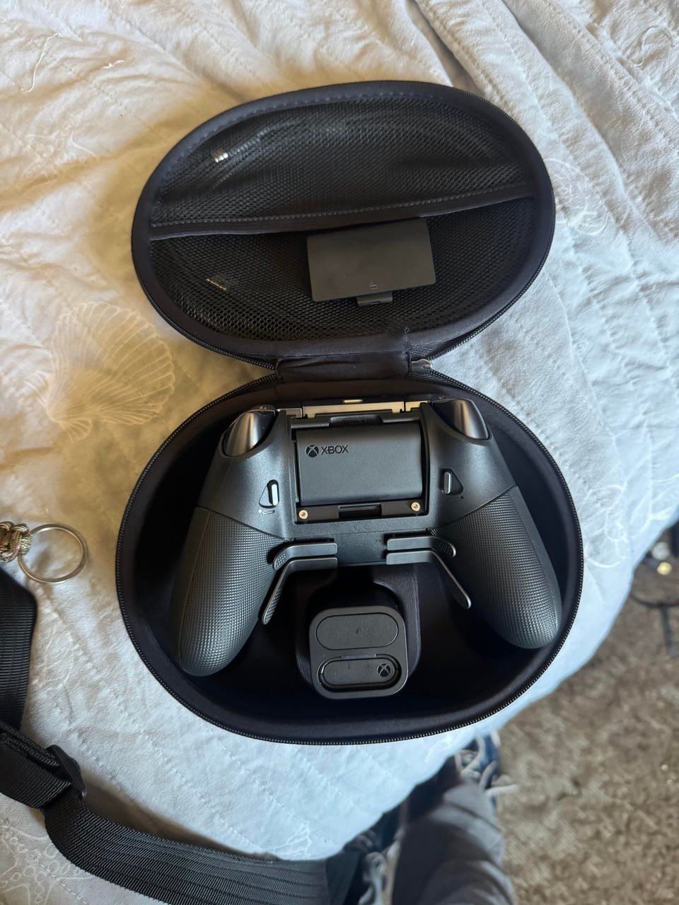 A black Xbox Elite controller rests inside a zippered, oval-shaped carrying case, positioned above a small charging dock on a textured white blanket.