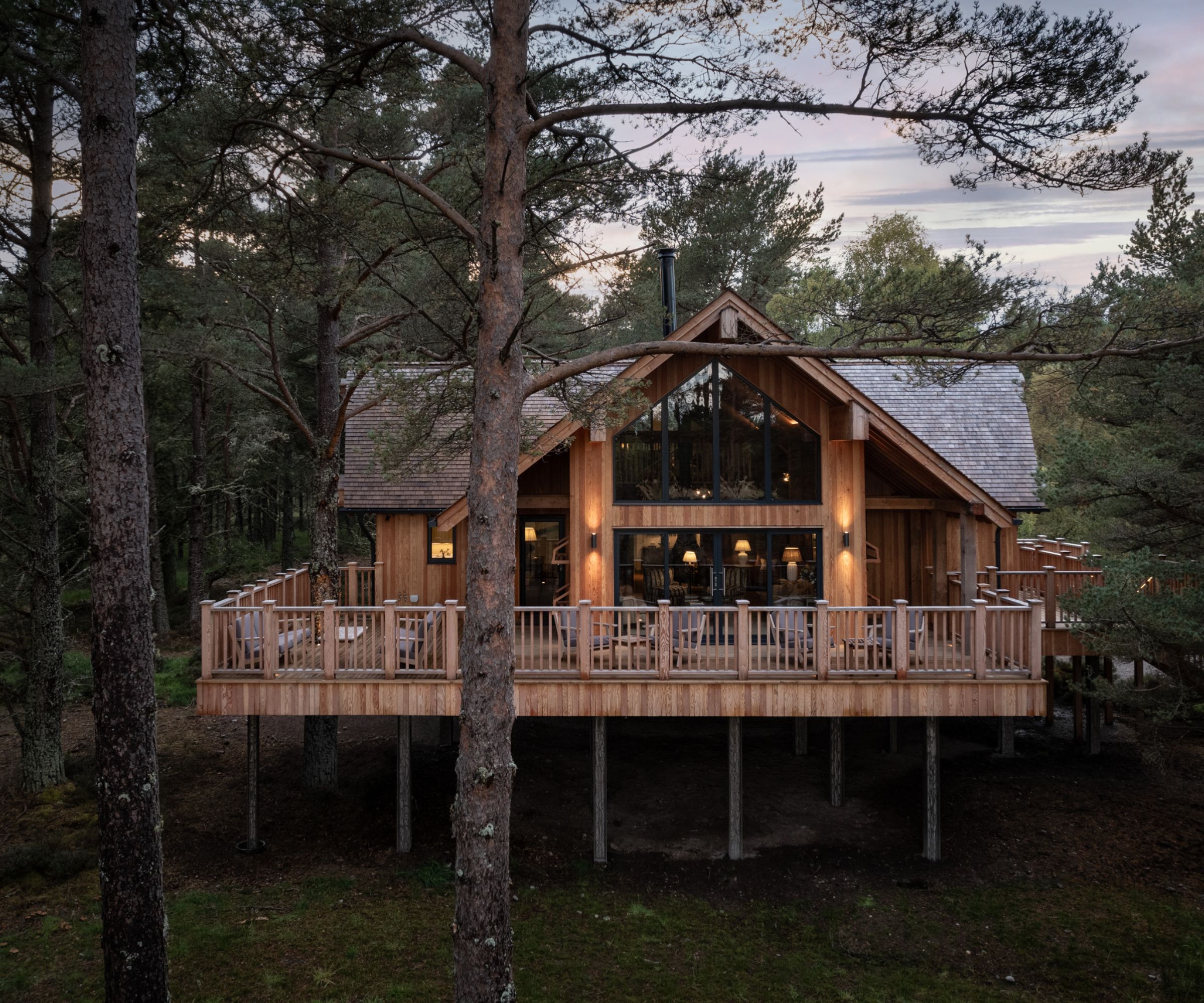 A luxury timber lodge elevated on stilts in a woodland setting, with a large wraparound deck and expansive glazed gable windows. The cabin blends beautifully into the pine forest surroundings, offering a cosy retreat immersed in nature.