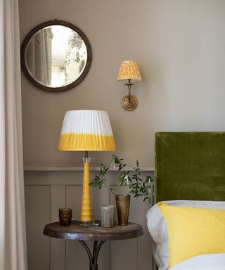 Yellow dip bedside lamp besides coordinating yellow pillowcase, and velvet green headboard, with mini yellow shade wall light, and round mirror on wall.