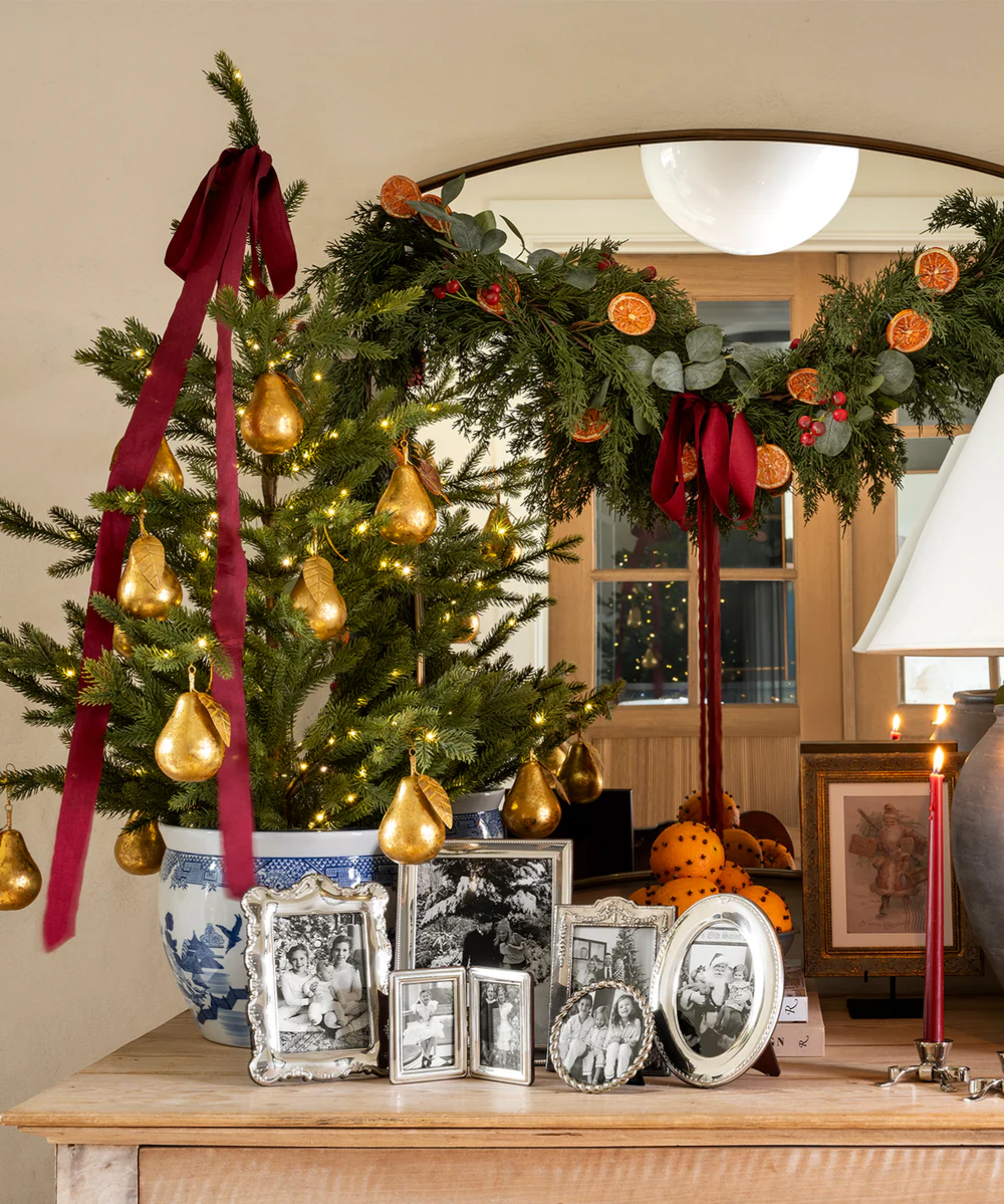 an entryway table styled for Christmas with a mini tree, framed photos, candles, and a mirror