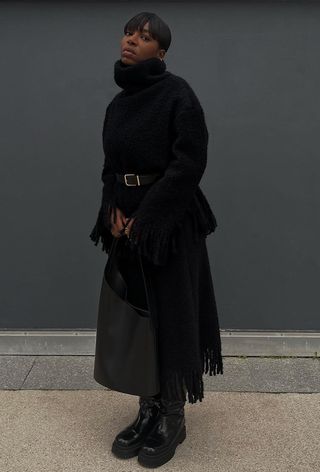 Black turtleneck outfit is shown in a photo of a French woman inside wearing a chunky black turtleneck sweater with a black belt ontop styled with a black knit fringe skirt, black tote bag, and black knee-high flat boots