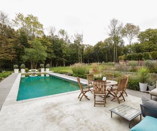 The view of a rectangular pool with pale hard landscaping with tiered planting nearby and. a circular outdoor dining table