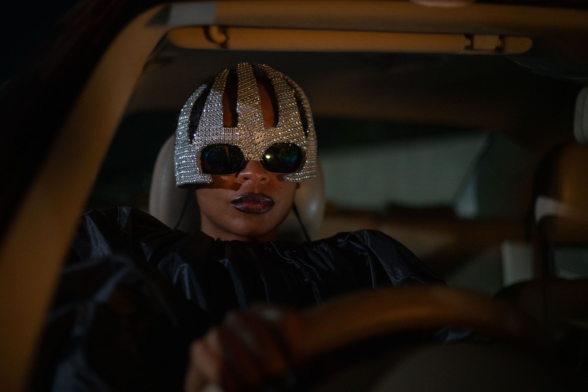 A woman (Susan Chardy as Shula) sits in the driver's seat of a car while wearing a bedazzled helmet, sunglasses, and a black trash bag, in 'On Becoming a Guinea Fowl.'