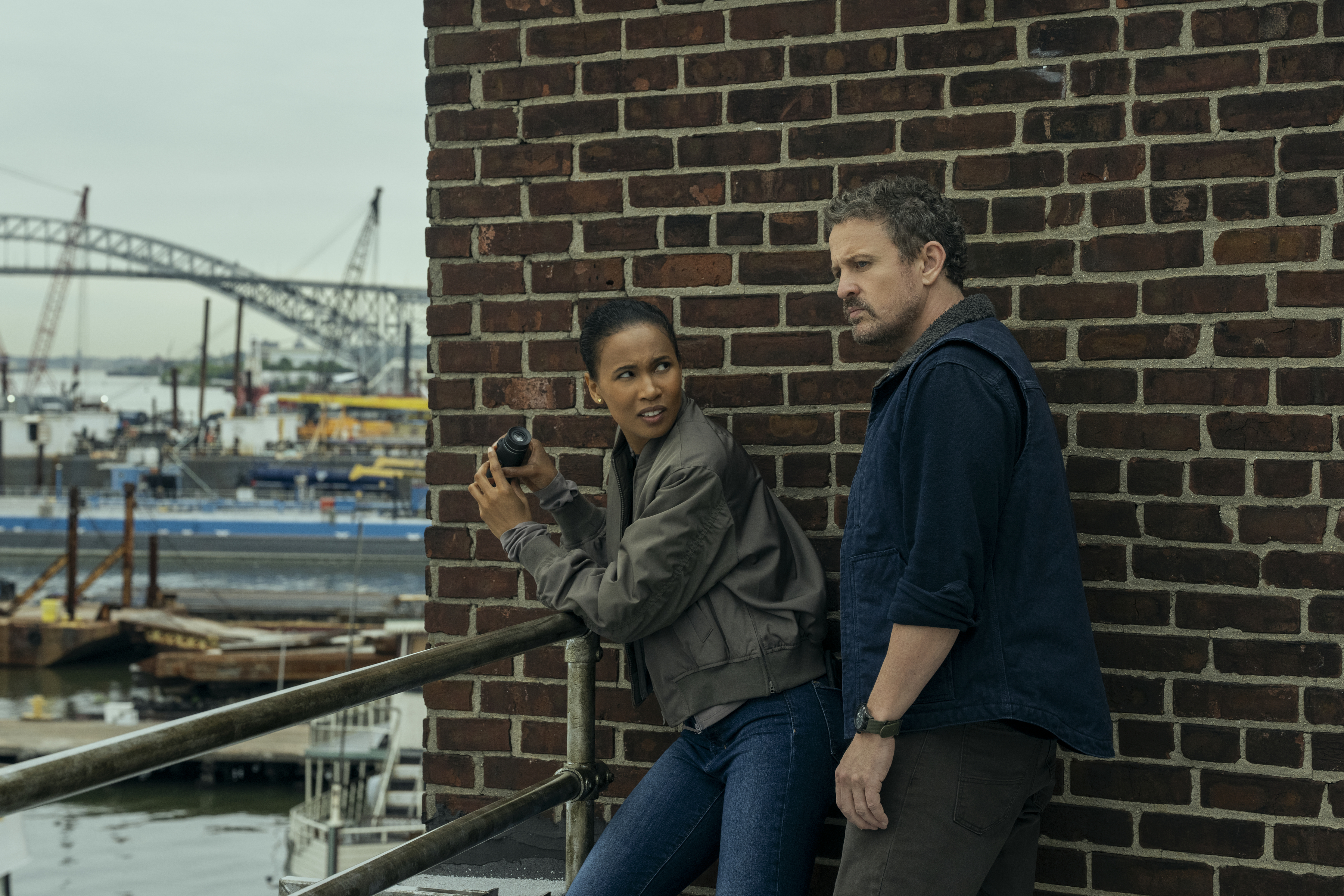Fola Evans-Akingbola as Chelsea Arrington looking over a navy yard with binoculars and David Lyons as Adam standing watch in the night agent season 3