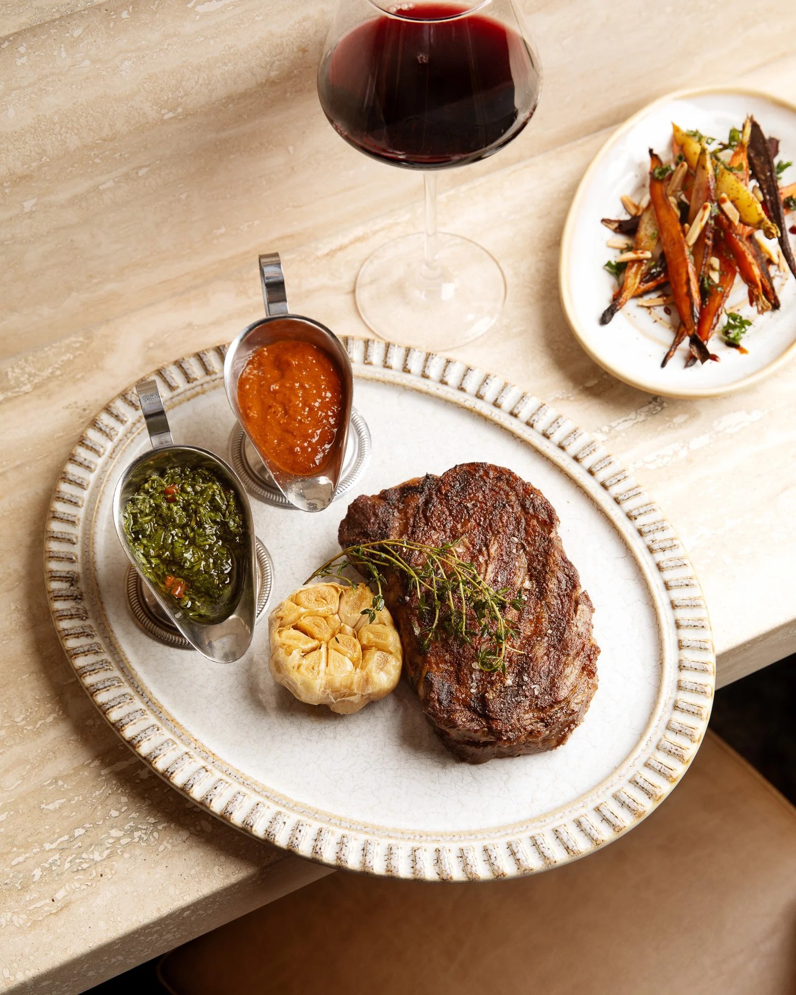 A pale wooden table is covered in food plates, including one with a golden-decorated, textural rim onto which sit a stake, a green and a red sauce in silver serving boats, respectively, and a garlic head, and another one filled with roast carrots.