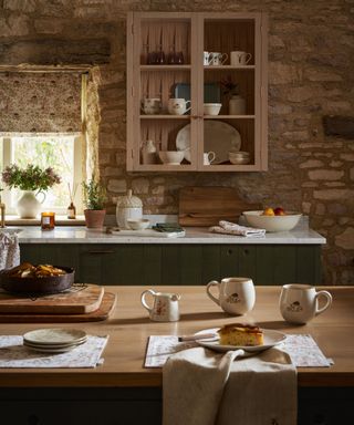 A kitchen with stone walls, a wooden cupboard and a wooden kitchen island with cutting boards, cups and dishes on it