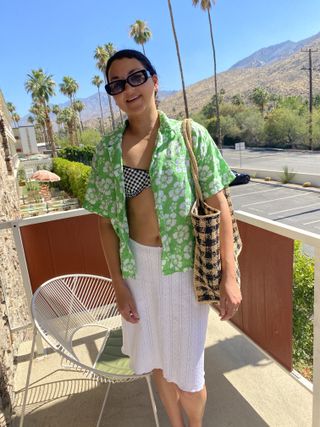 Woman on a balcony in Palm Springs wearing a floral shirt, white skirt, and straw tote.