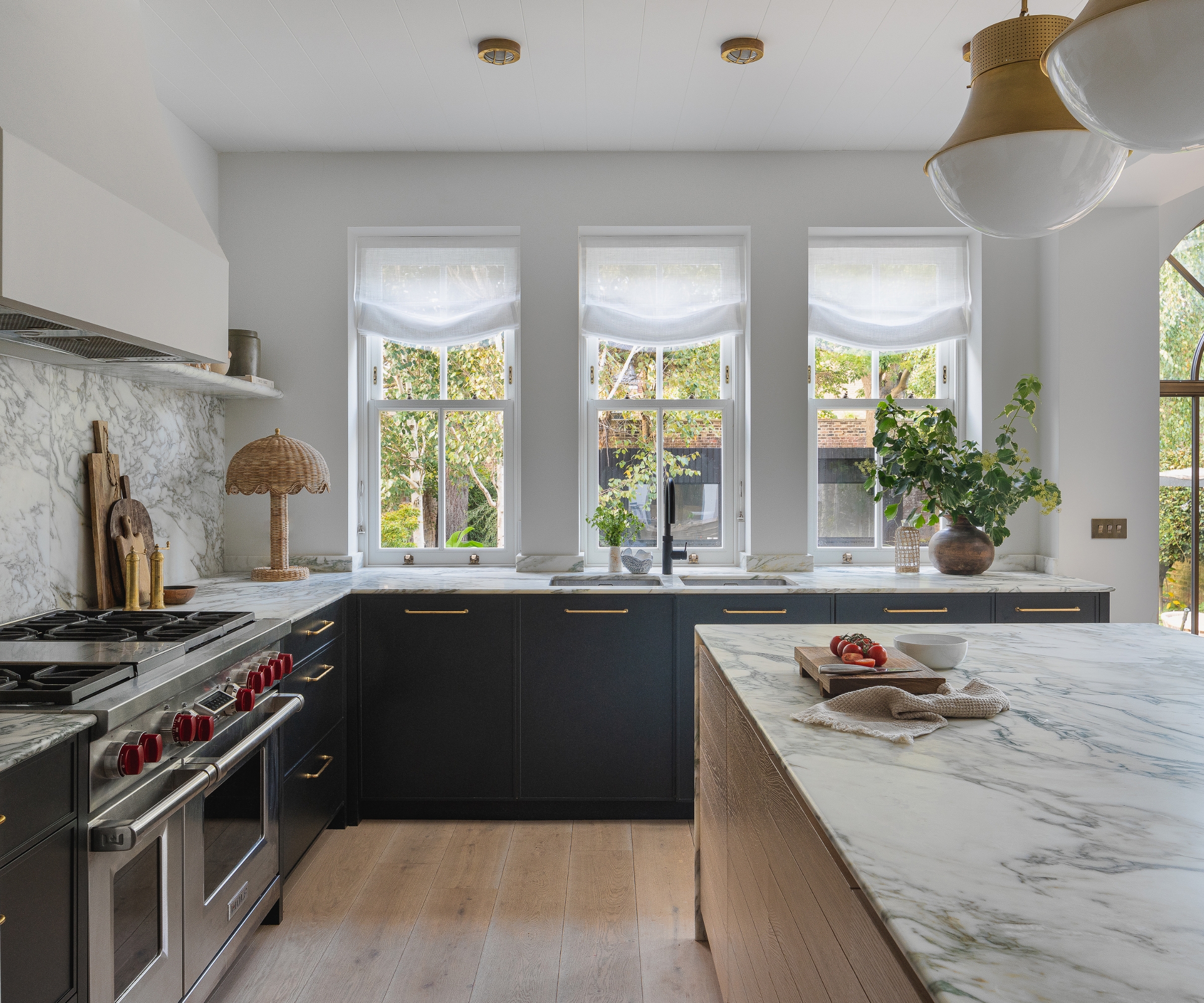 A navy and white kitchen with a wood island, marble countertops, sheer window treatments, and a rattan lamp