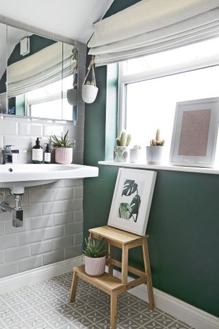 Bathroom with grey metro tiles on one wall, green paint on the other, grey patterned floor tiles, wall-hung sink and oak stepping stool with botanical artwork