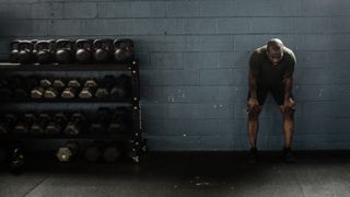 Man leaning against wall with hands on knees next to rack of kettlebells and dumbbells