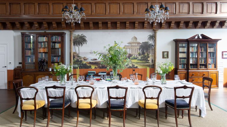 long formal dining table with white tablecloth and tall flowers with classical mural behind and wooden panelling and display cabinets with chandeliers above