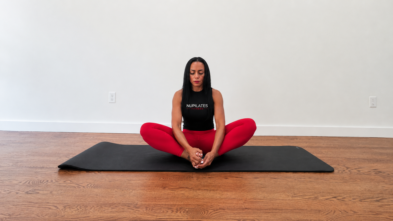Nicolle Lopez-Lamberty performs butterfly pose. She is sitting on the mat, with her feet together and knees out wide. Her hands clasp her ankles.