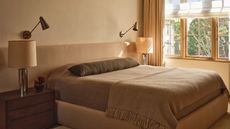 Image of a cozy but stylish bedroom drenched in a warm tan color. There is an extended, upholstered headboard, taupe bedding, and a dark bolster pillow.