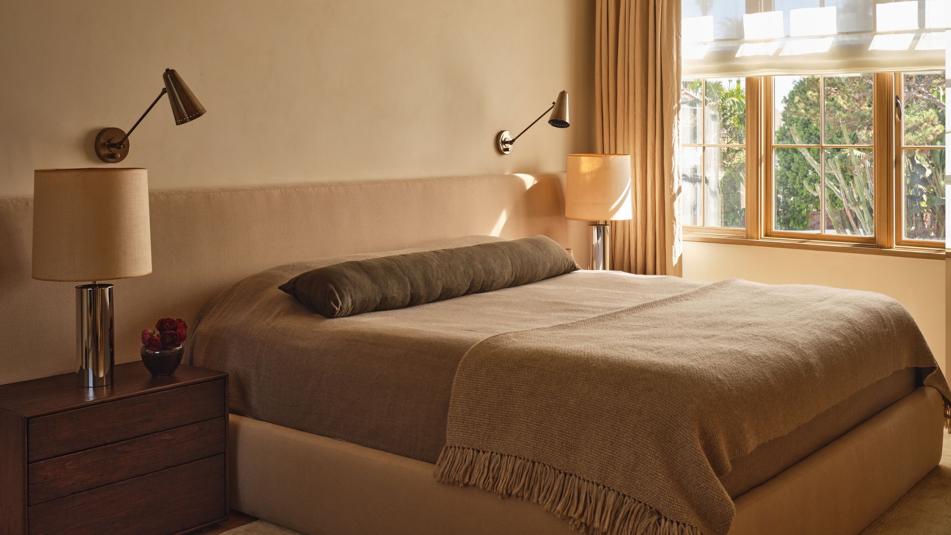 Image of a cozy but stylish bedroom drenched in a warm tan color. There is an extended, upholstered headboard, taupe bedding, and a dark bolster pillow.