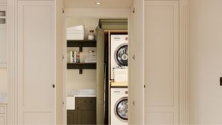 A hidden utility room tucked behind tall, panelled cabinet doors. Inside is a neatly stacked washer and dryer, shelving with laundry supplies, and olive green cabinetry with a farmhouse sink.