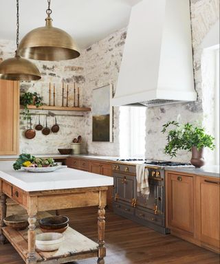 A kitchen with white washed stone walls and wooden cabinetry; a wooden kitchen island with a white countertop and a bowl of fruit in the center.