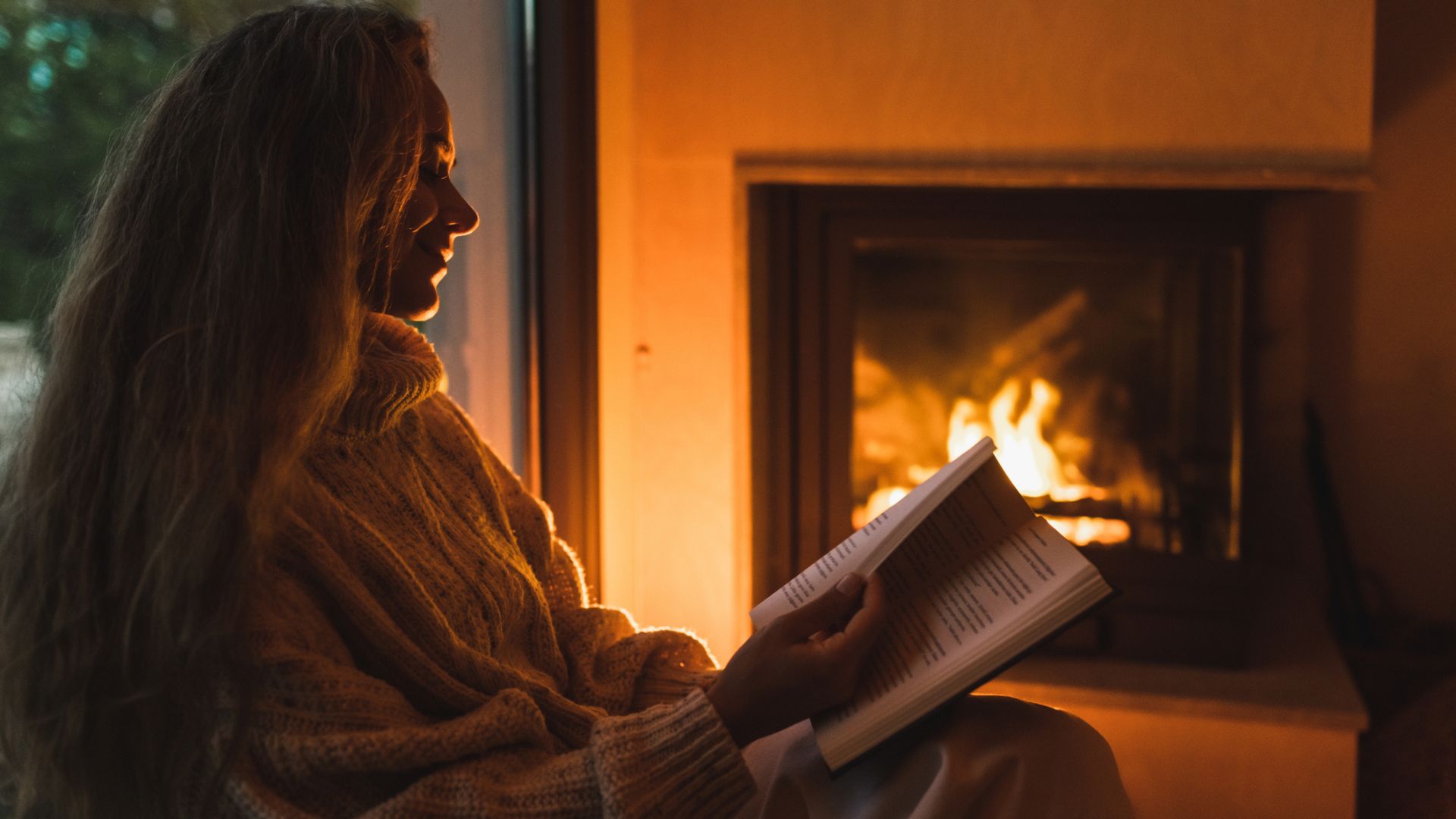 A woman wrapped in a blanket sat by a lit fireplace reading a book as the evening draws in