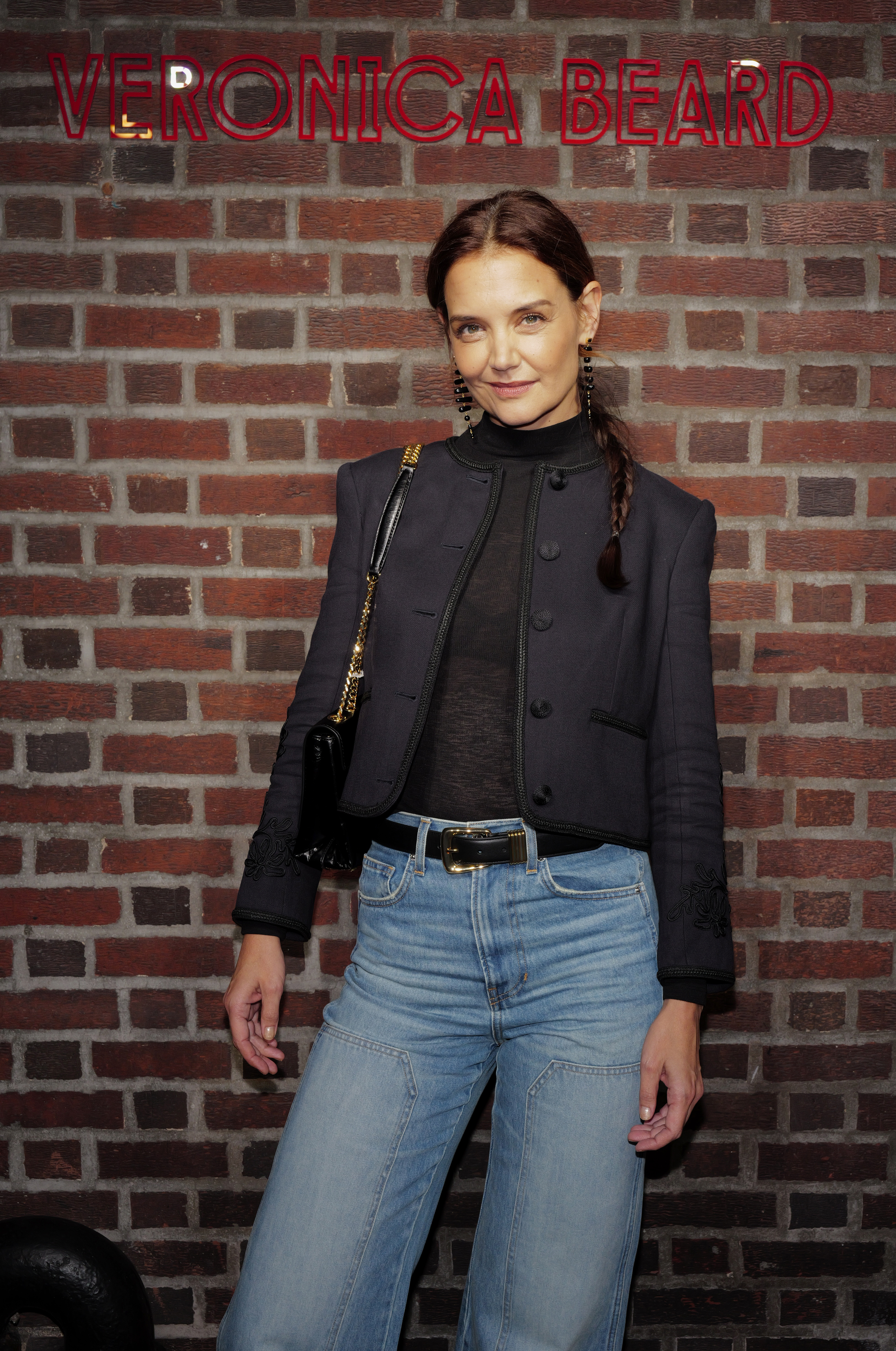a woman wearing a side braid, blue jeans, and a navy blue jacket taking a photo in front of a brick wall