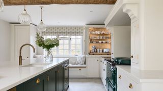 A country kitchen with a marble-topped island, brass tap, and dark green cabinetry. A cosy window seat, open shelving, and a classic range cooker add charm and warmth