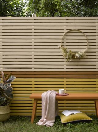 A yellow and white painted slatted fence in a backyard, with a bench in front of it with a pillow and a throw