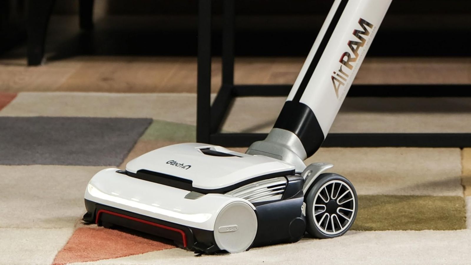 Gtech AirRAM 3 cordless vacuum cleaner on colorful, patterned rug in front of table legs.