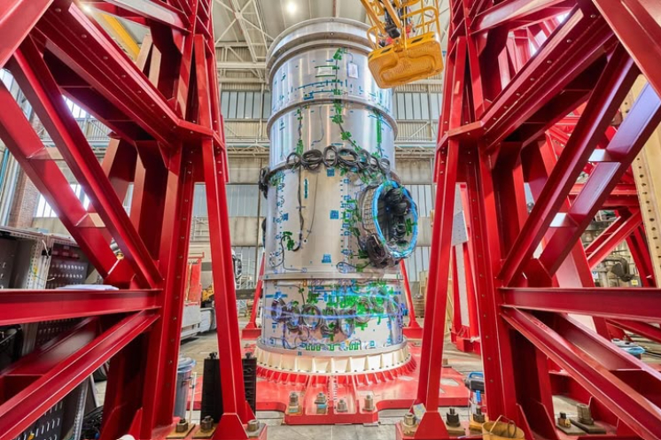 The cylindrical HALO module is seen standing vertically, surrounded by a red steel frame. The HALO module is silver and is lined with coils of green wires. ⁣