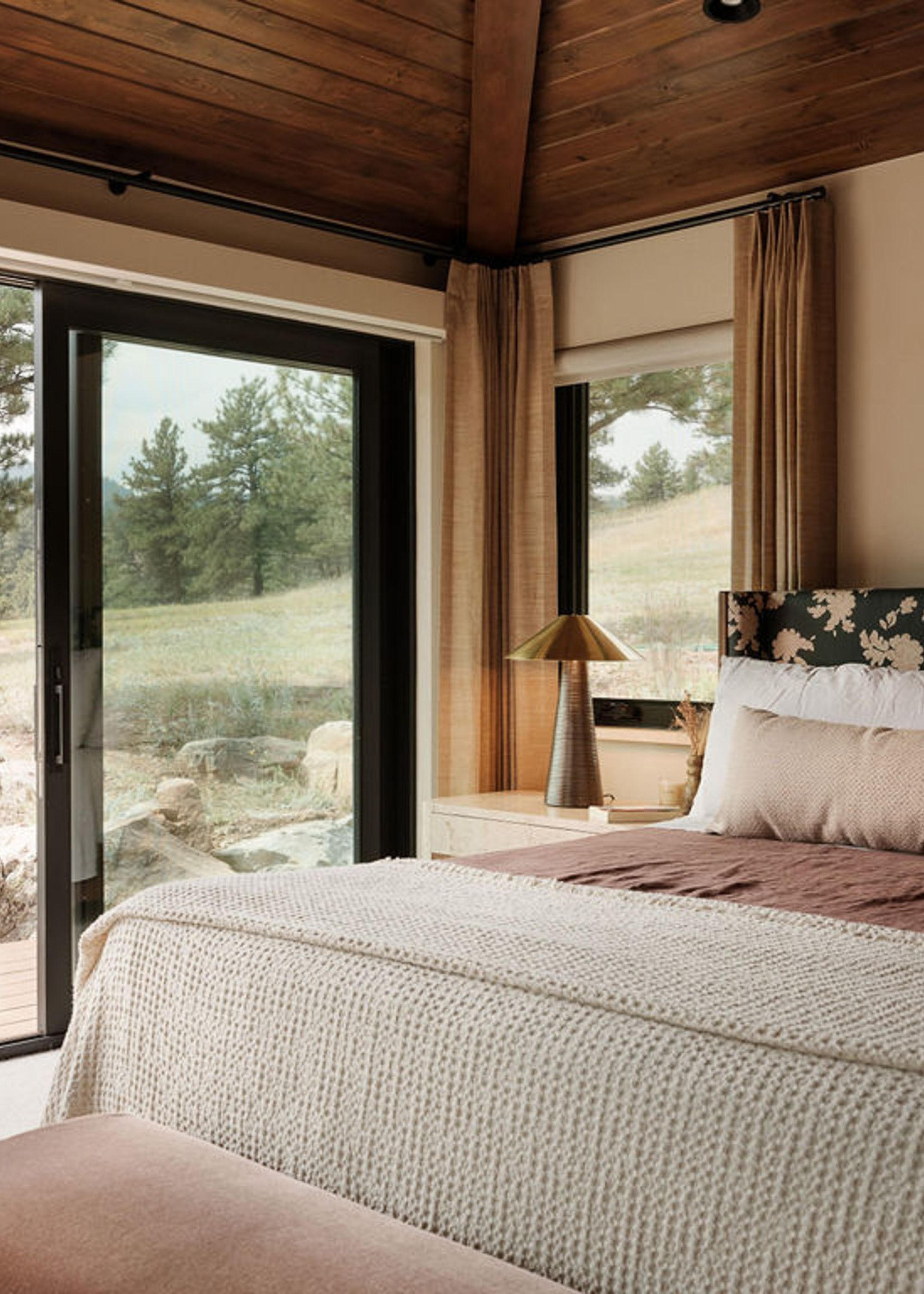 A bedroom with large windows and a door to the backyard. Also pictured is a bed with a patterned headboard, a brass lamp, and a textured coverlet