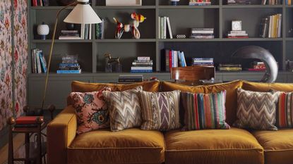 A maximalist living room with a mustard velvet sofa in front of built-in grey book shelves.