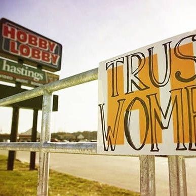 'Trust Women' sign posted on barrier in front of 'Hobby Lobby' and 'Hastings' signs