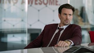 Chris Evans in a suit sitting at a desk in Pain Hustlers