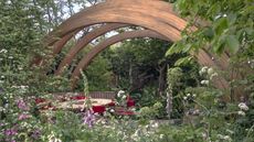 A show garden at Chelsea Flower Show with trees, shrubs, and flowering perennials under a large wooden arch