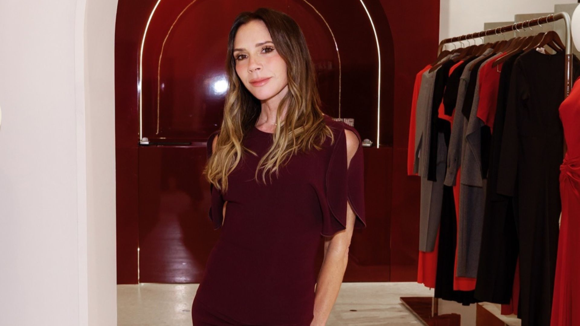 Victoria Beckham stands inside her stop wearing a burgundy dress with cream heels.