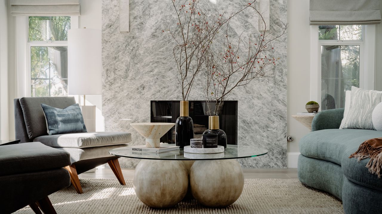 Colors that go with white marble: Living room with white walls, light wood floors, beige rug, sculptural coffee table, light teal sofa and large white marble fireplace