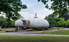 Exterior view of the light grey, curved 2014 Serpentine Pavilion by Smiljan Radic during the day. The structure sits on large stones and is surrounded by greenery